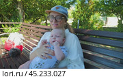Close-up of a woman in a baseball cap and glasses holding a baby in a patterned overalls. They are sitting on a park bench with lush green foliage in background. The camera moves gently into distance. Стоковое видео, видеограф Воловодов Владимир / Фотобанк Лори