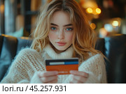 A young girl in a cozy sweater holds a bank card in her hands. Her gaze is directed at the camera, creating a sense of involvement and trust. The background is softly blurred, which emphasizes the cozy and comfortable environment, perhaps a cafe or a home space with soft light. This photo is great for illustrating themes related to online shopping, financial services or modern lifestyle. Стоковое AI-изображение, AI-художник Анна Кривицкая / Фотобанк Лори