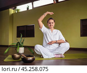A woman practices meditation in Kundalini yoga, sits in a comfortable lotus position on a mat in a room, in white clothes, with her hands showing the Chin Mudra gesture, full-length view. Стоковое фото, фотограф Сергей Чайко / Фотобанк Лори