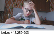 The boy solves math, does homework in the kitchen and yawns from boring tasks. The child writes the answer to the solution of the example in a notebook. The kid sits at the table and performs the task. Стоковое видео, видеограф Екатерина Золина / Фотобанк Лори