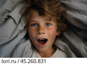 Top view, portrait of Boy with freckles lied in bed under blanket and yawns with wide opened mouth. He surprised, screaming or startled. Kid is surrounded by grey pillow and blanket. Стоковое AI-изображение, AI-художник Sergey Grishin / Фотобанк Лори