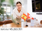 Positive young woman putting flag of Spain on the table with flag of Switzerland in conference room. Стоковое фото, фотограф Яков Филимонов / Фотобанк Лори