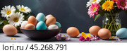 A bowl of colorful painted easter eggs on a table with first bright spring flowers. Concept of Pascha, Resurrection Sunday, Christian cultural holiday. Banner, copy space. Стоковое AI-изображение, AI-художник Sergey Grishin / Фотобанк Лори