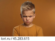 Portrait of disgruntled blond boy against orange background. The boy experiencing negative emotions due to fact that his family tore him away from computer games and forced him. Sulking Caucasian boy. Стоковое AI-изображение, AI-художник Sergey Grishin / Фотобанк Лори