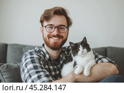 Young handsome smiling brunette man wearing glasses and checkered shirt bonding his tabby cat in hands on room background. Good friends. Friendship of pet and its owner. Cuddles. Copy space for text. Стоковое AI-изображение, AI-художник Марина Демидюк / Фотобанк Лори