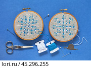 Top view on two hand embroidered cross stitch snowflakes in hoops on blue background and accessories for cross stitch. Winter crafts and Christmas gifts concept. Cross stitches as hobby. Flat lay, close-up. Стоковое фото, фотограф Виктория Катьянова / Фотобанк Лори
