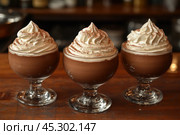 Three servings of chocolate mousse with whipped cream in glass bowls. The dessert is decorated with lush curls of cream and a light dusting of cocoa powder on top, creating an appetizing and aesthetic look. The background is blurred, which allows you to focus on the details of the dessert. This photo is perfect for illustrating a menu, culinary blogs, cafes or restaurants. Стоковое AI-изображение, AI-художник Анна Кривицкая / Фотобанк Лори