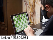 A man in a medical mask solves a chess problem online. Chess board with figures on a laptop screen. Leisure in self-isolation, in the twilight. International chess day. Стоковое фото, фотограф Vadzim Yakubovich / Фотобанк Лори