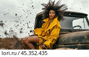 A model in a yellow raincoat poses dramatically on a vintage truck while mud splashes around her in an outdoor setting. Стоковое AI-изображение, AI-художник Сергей Толмачёв / Фотобанк Лори