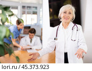 positive senior medical female colleague standing in meeting room and employees are talking behind her at table. Стоковое фото, фотограф Яков Филимонов / Фотобанк Лори
