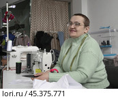 Женщина швея, с очень короткими волосами, после перенесённой болезни, в своей швейной мастерской сидит за швейной машинкой. A woman seamstress, with very short hair, after an illness, sits at a sewing machine in her sewing workshop. Стоковое фото, фотограф Наталья Пеньшина / Фотобанк Лори