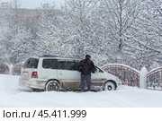 A man cleans the car from snow during a snowfall. Car parked in the snow. Lots of snow on the ground and trees. Cold snowy winter weather. Snowdrifts near the fence of the park. Стоковое фото, фотограф Андрей Степанов / Фотобанк Лори