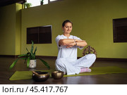 Woman practicing kundalini yoga, doing meditation sitting in lotus position on mat in the room, exercising in comfortable white clothes, full-length view. Стоковое фото, фотограф Сергей Чайко / Фотобанк Лори