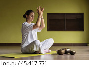 Woman practicing kundalini yoga, meditating sitting in comfortable position on mat in room and doing lotus mudra with hands up, full length view. Стоковое фото, фотограф Сергей Чайко / Фотобанк Лори