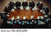 A business meeting at a large wooden table, around which men in business suits are sitting. The scene is shot from above, creating an emphasis on the shape of the table and the interaction of the participants. Documents and glasses of water are on the table, which emphasizes the formal atmosphere. Each participant is focused on the discussion, their gestures show involvement and seriousness. This image is ideal for illustrating business concepts, meetings, corporate culture, decision-making and teamwork. Стоковое AI-изображение, AI-художник Анна Кривицкая / Фотобанк Лори