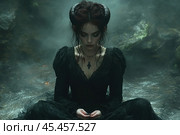 A young woman with a dark and mystical image. She is dressed in a black dress in the Gothic style, with lace elements. On her head are horns, which adds a demonic or mythological character to the image. The woman is sitting on the ground in a dark forest, surrounded by thick fog, creating an atmosphere of mystery and magic. Her gaze is lowered, and her hands are folded in a gesture of thought or meditation. The background is made in dark and muted tones, complementing the overall gothic and mystical theme. The photo is ideal for projects related to themes of fantasy, mysticism, gothic fashion, mythology and art photography. Gothic. Стоковое AI-изображение, AI-художник Анна Кривицкая / Фотобанк Лори