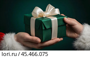 Hands of Santa Claus in close-up with a gift box in his hands on a green background. Gifts from Santa Claus. Стоковое AI-изображение, AI-художник Photojuli / Фотобанк Лори