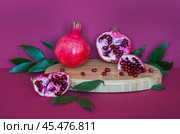 Beautiful still life with ripe pomegranates and pomegranate tree leaves on a wooden podium on a burgundy background. Advertisement of natural organic seasonal fruits with a high content of vitamins. Стоковое фото, фотограф Виктория Катьянова / Фотобанк Лори
