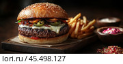 An appetizing burger with a beef patty and crispy bacon, fresh lettuce and tomatoes, covered with a golden brioche bun in a perfect wrapper fresh fried French fries in the background. Стоковое AI-изображение, AI-художник Ярославский Максим / Фотобанк Лори