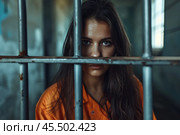 A female inmate in an orange jumpsuit stands behind metal bars, looking directly at the camera. Her expression reflects confinement in a prison cell. Стоковое AI-изображение, AI-художник Sergey Grishin / Фотобанк Лори