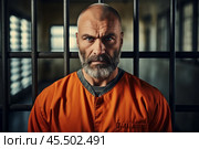 A prisoner in an aged orange uniform stands behind metal bars, looking intently at the camera. This portrayal captures the stark reality of life in confinement within a detention center. Стоковое AI-изображение, AI-художник Sergey Grishin / Фотобанк Лори