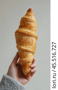 Hand holding a freshly baked croissant with golden layers in a minimalistic setting. Стоковое AI-изображение, AI-художник Юлия Гапеенко / Фотобанк Лори