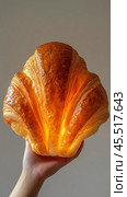 Hand holding a freshly baked croissant with golden layers in a minimalistic setting. Стоковое AI-изображение, AI-художник Юлия Гапеенко / Фотобанк Лори