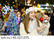 Girl in red Santa hat and with Christmas tree toys in her hands smiles dreamily. Стоковое фото, фотограф Яков Филимонов / Фотобанк Лори