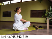 Woman practicing kundalini yoga, doing twisting exercises while sitting in lotus position, practicing in room in white clothes on mat. Стоковое фото, фотограф Сергей Чайко / Фотобанк Лори