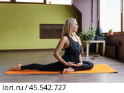 Athletic middle-aged blonde woman leading a healthy lifestyle does the Rajakapotasana exercise, the first variation of the pigeon pose, trains in sportswear on a mat in a yoga studio, full-length view. Стоковое фото, фотограф Сергей Чайко / Фотобанк Лори