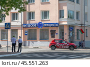 Город Витебск, Беларусь. 1-я улица Доватора. Красная легковая  машина с надписью на боту "Coca-Cola" едет по улице мимо продовольственного магазина "Витебские продукты". На углу стоят спиной к зрителю два милиционера (2024 год). Редакционное фото, фотограф Наталья Николаева / Фотобанк Лори