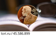 Moscow December 03 2024. Alfred Nobel's image on gold coin on physics book and reading glasses in background. Nobel Prize award. Nobel Prize for Literature concept. Редакционное фото, фотограф Скрипачев Антон / Фотобанк Лори