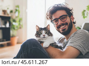 Young handsome smiling brunette man wearing glasses and checkered shirt bonding his tabby cat in hands on room background. Good friends. Friendship of pet and its owner. Cuddles. Copy space for text. Стоковое AI-изображение, AI-художник Марина Демидюк / Фотобанк Лори