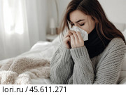 Young sick woman with cold or flue sitting on grey couch, holding a tissue to clear, blowing runny nose. Sneezing Girl under gray plaid blanket with handkerchief to dripping nose, blowing snot at home. Стоковое AI-изображение, AI-художник Марина Демидюк / Фотобанк Лори