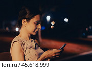 Young woman using phone while sitting on bench at night city street. Cinematic film look photo with girl chatting or messaging in her smartphone. Стоковое фото, фотограф Константин Григорьев / Фотобанк Лори