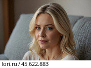 Head shot portrait pretty blonde woman looking at camera, sits alone on sofa photoshooting, enjoy pleasant pastime weekend at home at own or rented apartment. Beautiful mature female profile picture. Стоковое AI-изображение, AI-художник Александр Поташев / Фотобанк Лори