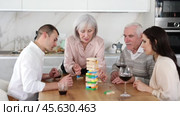 Elderly woman engrossed in playing jenga with husband, adult son and daughter-in-law during family gathering with wine at home, concentrating on extracting block without collapsing tower. Стоковое видео, видеограф Яков Филимонов / Фотобанк Лори