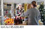 Cheerful female store clerk in striped jumper, maroon apron and shiny Santa hat helping customer choose bottle of alcoholic drink for Christmas celebration at small, holiday-decorated grocery shop. Стоковое видео, видеограф Яков Филимонов / Фотобанк Лори