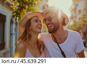 A couple shares a lighthearted moment, laughing together in a sunlit city street, wearing stylish hats and sunglasses, surrounded by colorful buildings. Стоковое AI-изображение, AI-художник Sergey Grishin / Фотобанк Лори