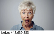 An elderly woman shows a moment of surprise, her eyes wide and mouth open in excitement. She wears a light blue shirt and stands against a soft, neutral background. Стоковое AI-изображение, AI-художник Sergey Grishin / Фотобанк Лори