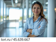 A Hispanic female facility manager smiles warmly while standing with her arms crossed in a contemporary building filled with natural light, showcasing her professionalism and confidence. Стоковое AI-изображение, AI-художник Sergey Grishin / Фотобанк Лори