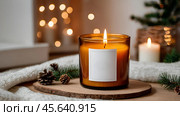 A mock-up of a cozy burning candle made of amber glass with an empty white label in a festive, New Year's home interior. Стоковое AI-изображение, AI-художник Photojuli / Фотобанк Лори