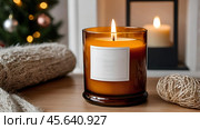 A mock-up of a cozy burning candle made of amber glass with an empty white label in a festive, New Year's home interior. Стоковое AI-изображение, AI-художник Photojuli / Фотобанк Лори