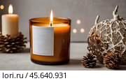 A mock-up of a cozy burning candle made of amber glass with an empty white label in a festive, New Year's home interior. Стоковое AI-изображение, AI-художник Photojuli / Фотобанк Лори