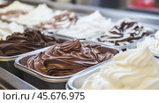 Chocolate, vanilla, strawberry ice cream tubs. Freshly churned various ice cream in a metal containers. Freezer full of icecream in the boxes in cooled counter. Gelato display of many flavors. Стоковое AI-изображение, AI-художник Марина Демидюк / Фотобанк Лори