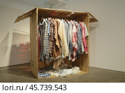 Cardboard box used as makeshift closet filled with casual shirts and clothes. Стоковое AI-изображение, AI-художник Иван Рябоконь / Фотобанк Лори