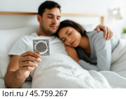 Пара лёжа в постели держит блистер презерватива. Стоковое AI-изображение, AI-художник Anna Leeva / Фотобанк Лори