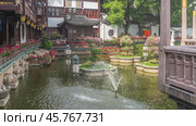 A fragment of a Chinese park. A pond with decorative flower beds, flowering bushes, and a fountain. Old traditional buildings with curved tiled roofs, ornaments around the perimeter. Shanghai. Стоковое фото, фотограф Вера Ивановна Кубарева / Фотобанк Лори