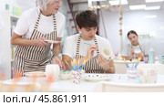 Experienced mature female ceramicist teaching young enthusiastic guy art of ceramic glazing and painting in modern pottery studio. Стоковое видео, видеограф Яков Филимонов / Фотобанк Лори