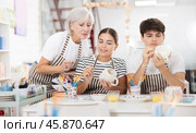 Mature female ceramicist teaching young couple to paint pottery in studio. Стоковое фото, фотограф Яков Филимонов / Фотобанк Лори